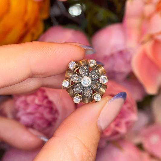 A close-up of a hand holding an antique floral ring with a gold-toned filigree pattern and clear diamonds, set against a soft-focus background of pink flowers.