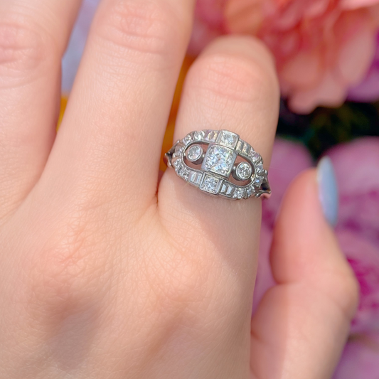 A close-up of a hand wearing a white gold Art Deco style ring with diamond gemstones, set against a soft-focus background of pink flowers.