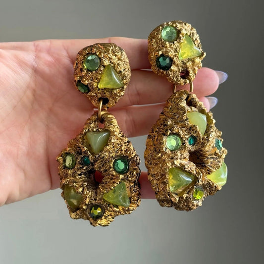A pair of chunky, textured gold statement drop earrings decorated with green and light yellow stones, held up by a hand against a neutral gray background.