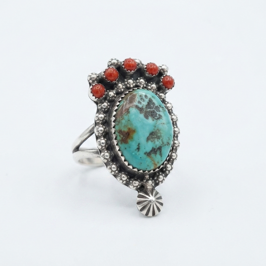 A sterling silver ring featuring a large oval turquoise cabochon with brown matrix patterns, accented by six small round red coral stones along the top, set against a plain white background.