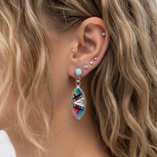 A close-up of a woman's ear wearing a silver-toned dangle earring with a colorful geometric inlay and several small ear studs, against a light neutral background.