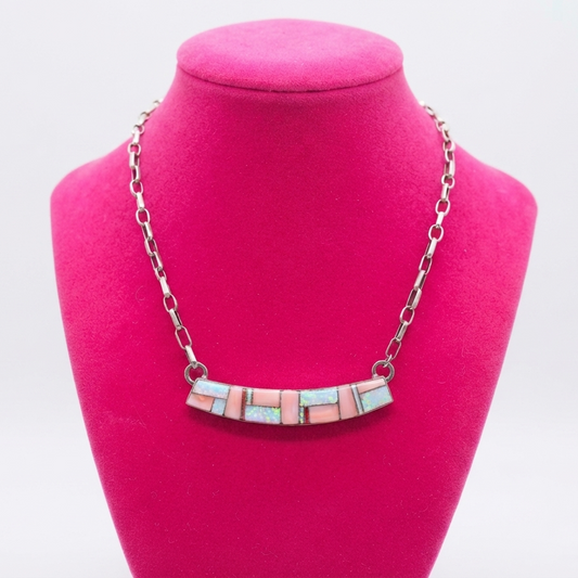 A silver chain necklace with a bar-shaped centerpiece made of small rectangular pink and light blue stones, displayed on a bright pink jewelry bust against a white background.