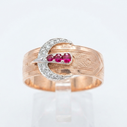 A gold ring with a buckle design, featuring a row of diamond stones and three pinkish-red ruby gemstones in the center, displayed on a clear stand against a white background.