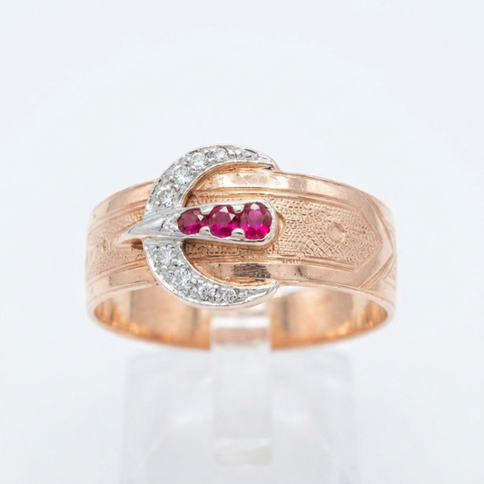A gold ring with a buckle design, featuring a row of diamond stones and three pinkish-red ruby gemstones in the center, displayed on a clear stand against a white background.