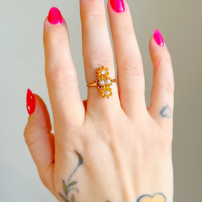 A close-up of a hand with bright pink and red nail polish, wearing a gold ring with a floral setting and four diamonds, set against a plain light gray background.