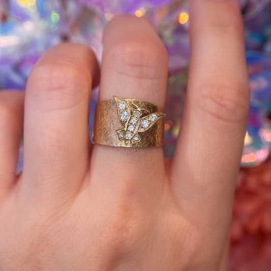 A gold ring with a textured band and a butterfly design paved with small clear crystals, shown worn on a finger against a colorful, iridescent bokeh background.