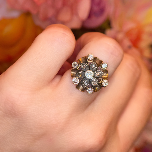 A close-up of a hand wearing an antique floral ring with clear diamond gemstones and a gold setting, set against a soft-focus background of pink and orange flowers.