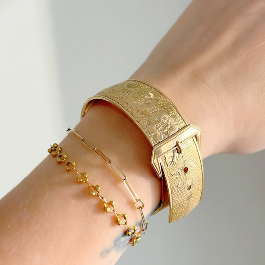 A close-up of a person's wrist wearing a wide textured gold cuff bracelet alongside a delicate gold chain bracelet with small floral charms.