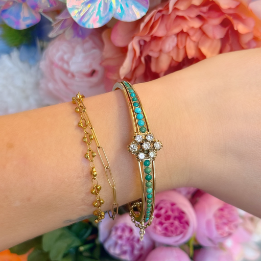 A close-up of a person's wrist wearing two gold bracelets, one with turquoise and clear diamond stones, set against a background of colorful flowers.