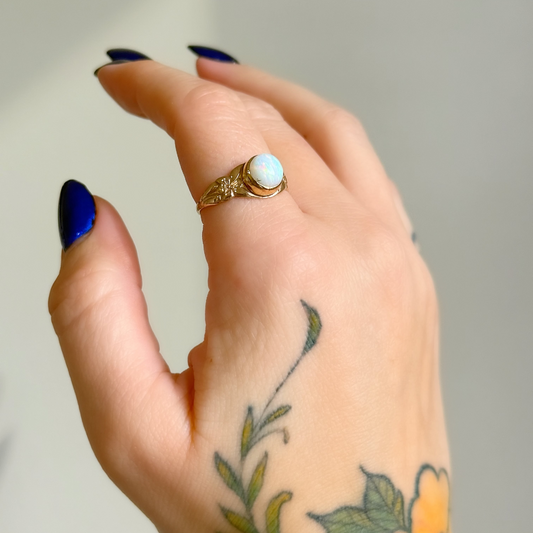 A close-up of a hand with blue nail polish and a floral tattoo, wearing a gold ring with a round opal cabochon stone.