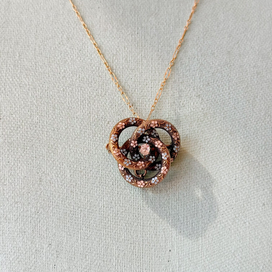 A gold necklace with a double-knot interlocking pendant set with small clear diamond stone, surrounded by tiny flowers, displayed on a textured off-white fabric background.