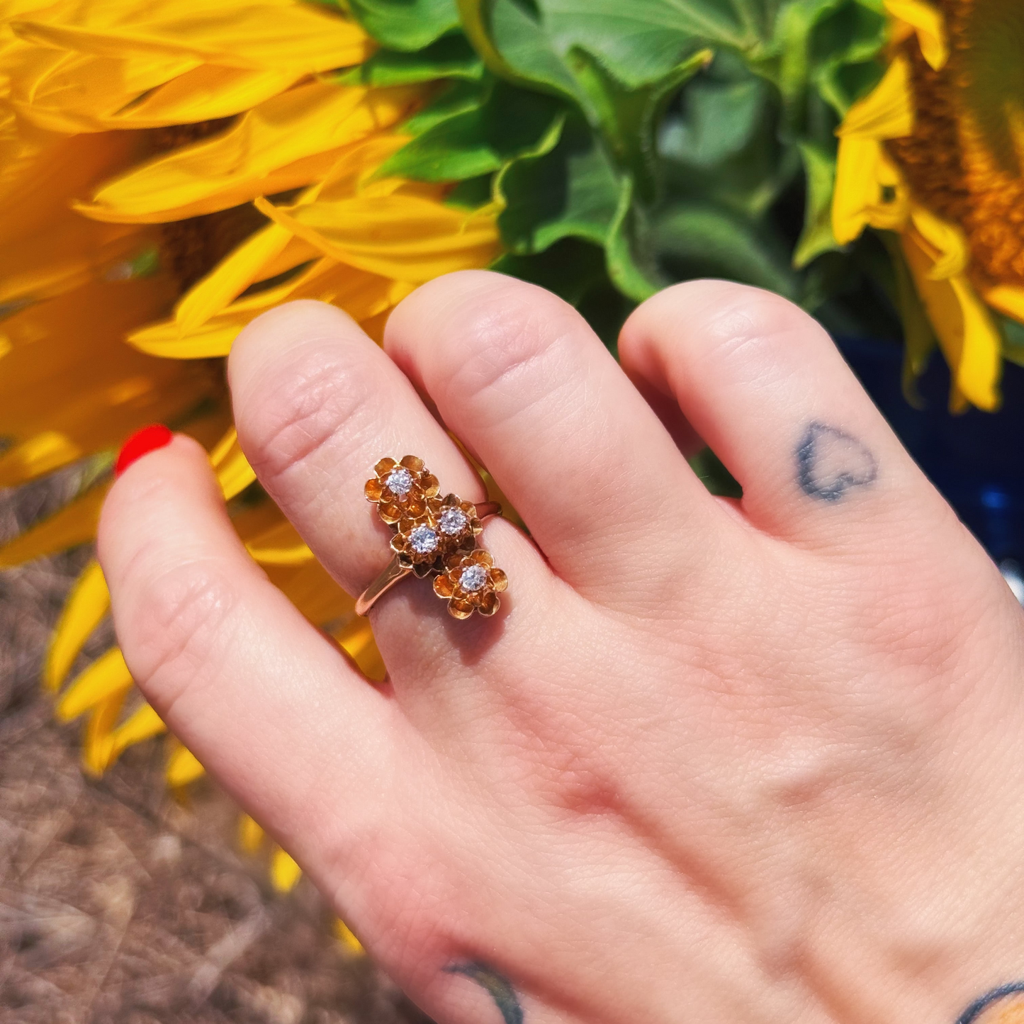 A close-up of a hand wearing a gold ring with three small clear diamond gemstones, set against a background of yellow sunflowers.