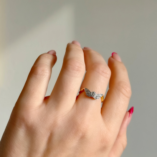 A close-up of a hand wearing a silver ring with a textured, organic pattern and small clear stones, set against a soft, neutral gray background.