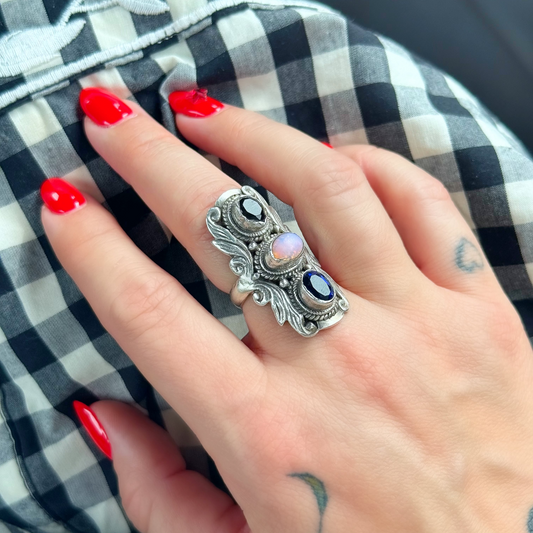 A close-up of a hand with red nail polish wearing a silver-toned ring with three oval gemstones, resting on a black and white gingham fabric surface.