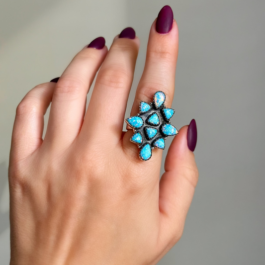 A close-up of a hand with dark purple manicured nails wearing a silver ring set with multiple bright turquoise blue teardrop-shaped opal stones.