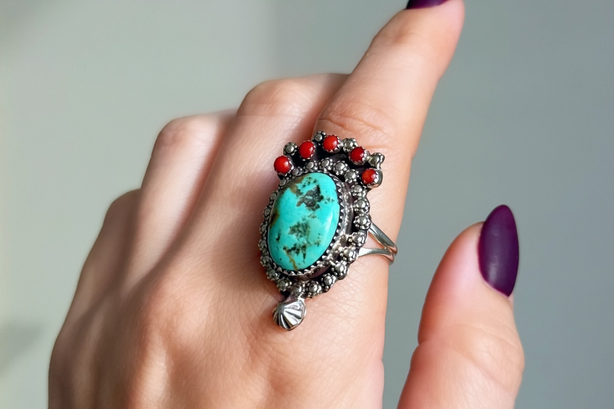 A close-up of a hand with dark purple nail polish wearing a silver ring featuring a large oval turquoise stone and small red coral stones along the top.