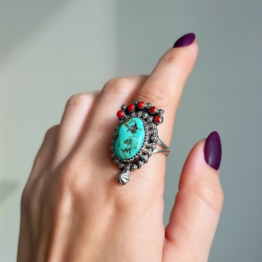 A close-up of a hand with dark purple nail polish wearing a silver ring featuring a large oval turquoise stone and small red coral stones along the top.