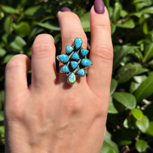 A close-up of a hand wearing a silver ring set with multiple round turquoise-blue opal stones, photographed outdoors against a background of green leaves.