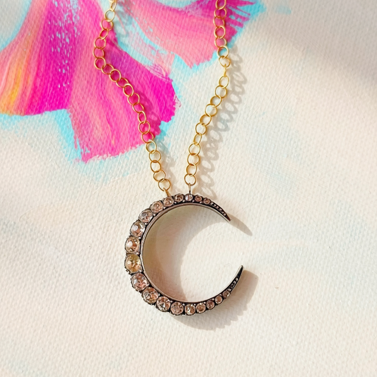 A gold chain necklace with a crescent moon pendant adorned with small clear rhinestones, displayed on a textured white surface with a colorful paint stroke in the background.