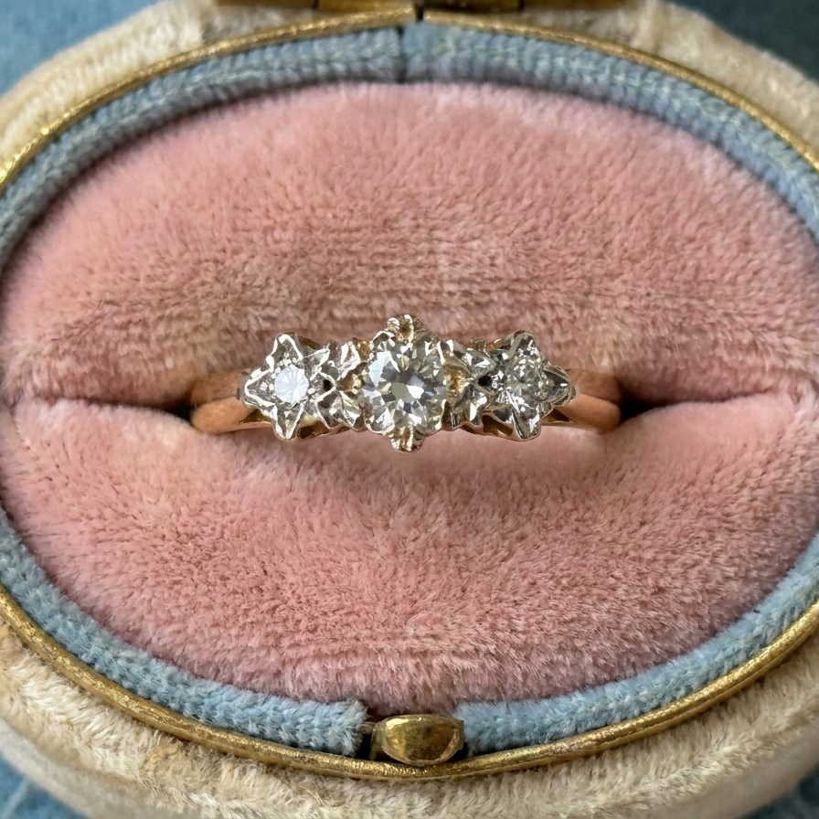 A three-stone diamond ring with a gold band, displayed in an open vintage-style ring box with a pink velvet cushion and blue-gray fabric lining.