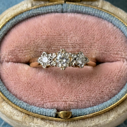 A three-stone diamond ring with a gold band, displayed in an open vintage-style ring box with a pink velvet cushion and blue-gray fabric lining.