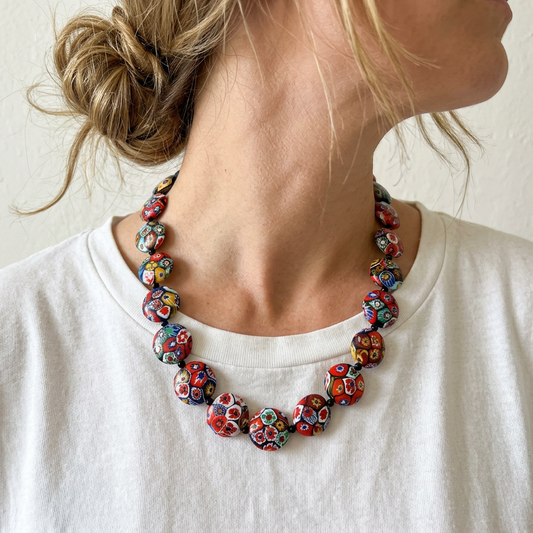 A close-up lifestyle shot of a woman wearing a chunky glass bead necklace with a vibrant multicolor pattern of red, blue, yellow, and white, paired with a white t-shirt.