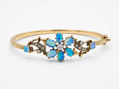 A gold hinged bangle bracelet featuring a central floral arrangement of blue opal gemstones and small clear diamond stones, set against a plain white background.