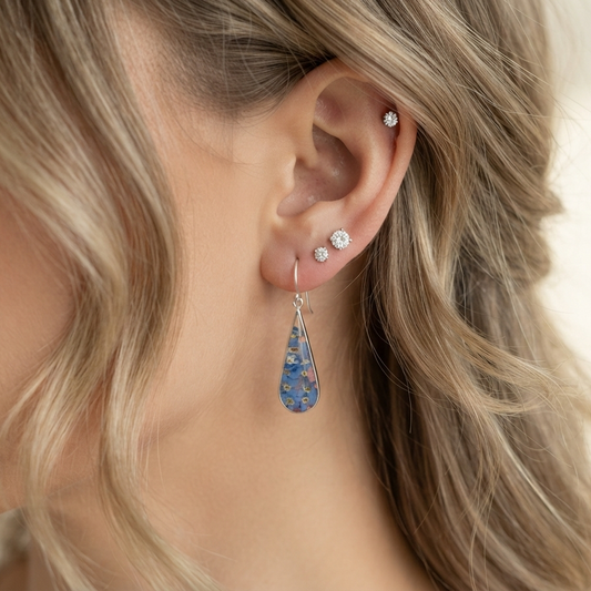 A close-up of a woman's ear wearing a dangling teardrop earring with a blue floral pattern and silver-toned studs.