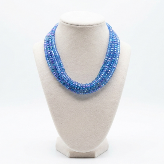 A multi-strand beaded necklace in shades of blue and purple, displayed on a cream-colored fabric jewelry bust against a white background.