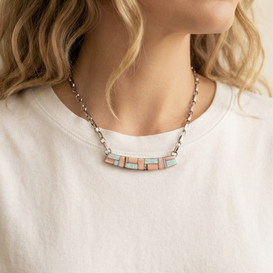 A close-up of a woman wearing a silver-toned necklace with a curved bar of rectangular pastel tiles in pink, peach, and mint green, paired with a white crewneck t-shirt.