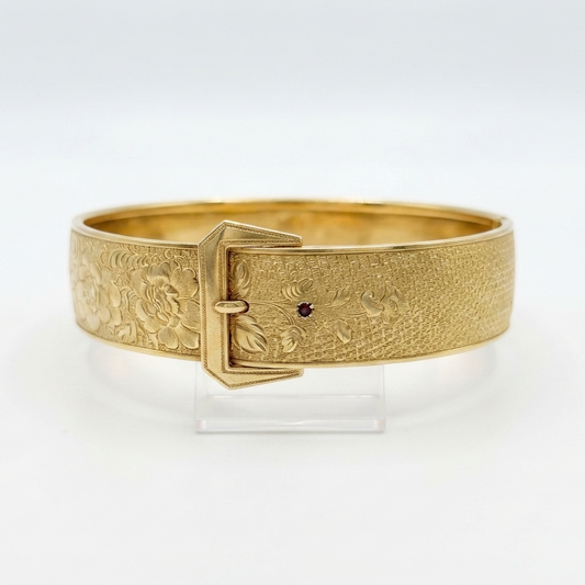 A gold-colored metal buckle bangle bracelet with an embossed floral pattern, displayed on a clear acrylic stand against a white background.