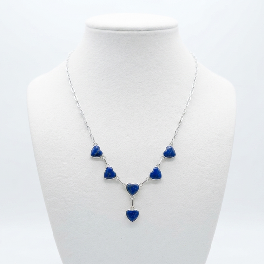 A silver necklace with five small blue heart charms and one larger dangling blue heart, displayed on a white jewelry bust against a light gray background.