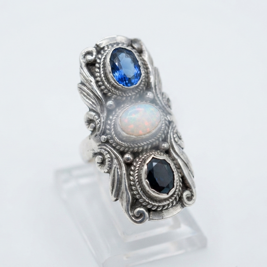A silver ring with three oval gemstones (blue sapphire, white opal, and dark blue/black stone) displayed on a clear acrylic stand against a white background.