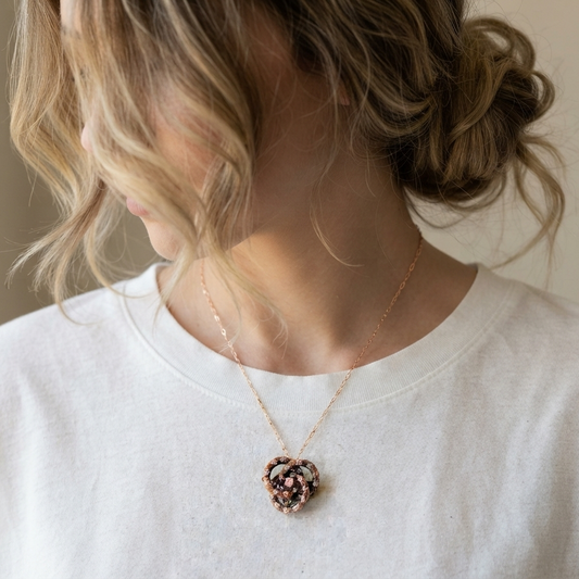 A close-up lifestyle shot of a woman wearing a delicate gold chain necklace with a knot-shaped woven pendant enamel flowers and a diamond, paired with a white crewneck t-shirt.