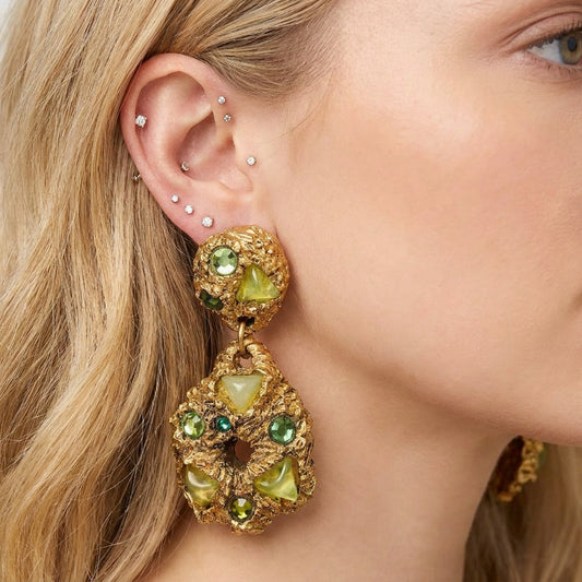 Close-up of a woman wearing gold earrings with green gemstones on a plain background